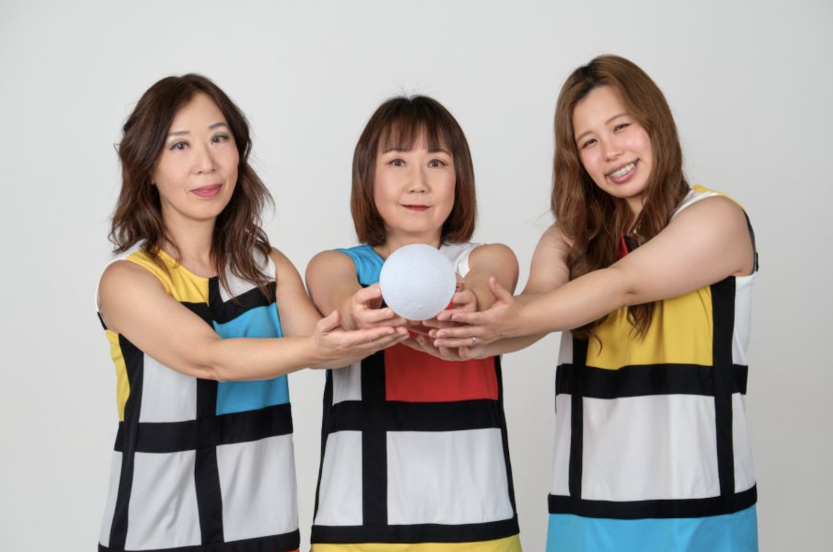 Pop-punk band Shonen Knife's three members wear matching Mondrian-style dresses and hold a white sphere together against a plain background.